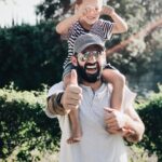 Smiling dad giving a thumbs up gesture while his son sits happily on his shoulders, creating a heartwarming moment of fatherly joy and bonding.