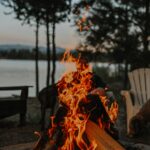 Flames flicker and dance in an outdoor fire pit situated by the serene edge of a tranquil lake. The warm glow of the fire contrasts against the cool, reflective surface of the water, creating a picturesque scene of natural beauty and relaxation.