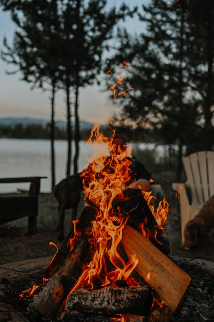 Flames flicker and dance in an outdoor fire pit situated by the serene edge of a tranquil lake. The warm glow of the fire contrasts against the cool, reflective surface of the water, creating a picturesque scene of natural beauty and relaxation.