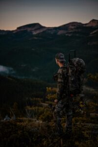 A bow hunter stands at dusk on the edge of a mountain, silhouetted against the fading light. With bow in hand, he gazes outwards, surveying the majestic landscape spread before him. The hunter's figure is outlined against the dusky sky, evoking a sense of anticipation and connection with the wilderness