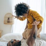 A heartwarming scene captured as a man lies on a bed, joyfully tossing his young son playfully into the air. Both father and child wear bright smiles, radiating happiness and bonding in a moment of carefree playfulness and affection